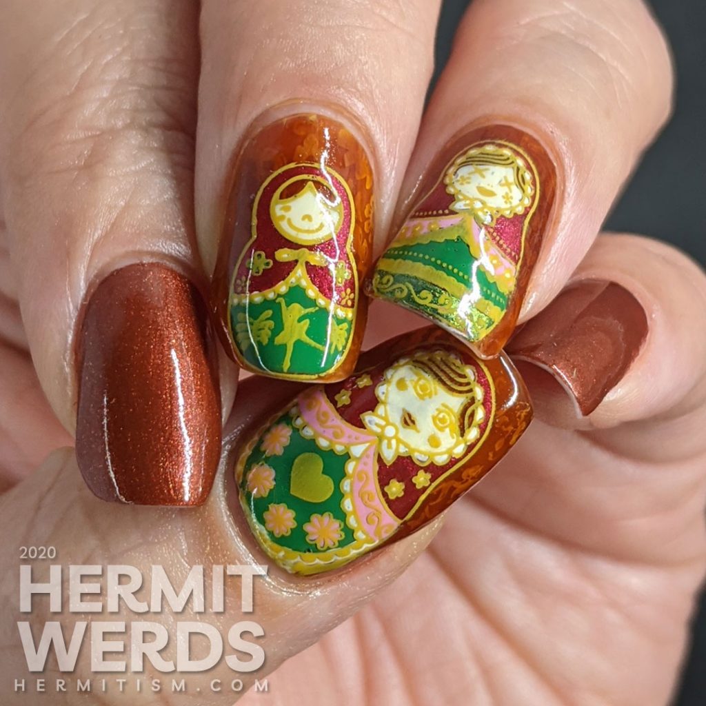 Nail art with stamping decals of beautiful Russian nesting dolls or matryoshka dolls in pinks, reds, and greens on a rusty red background with subtle jelly sandwich.