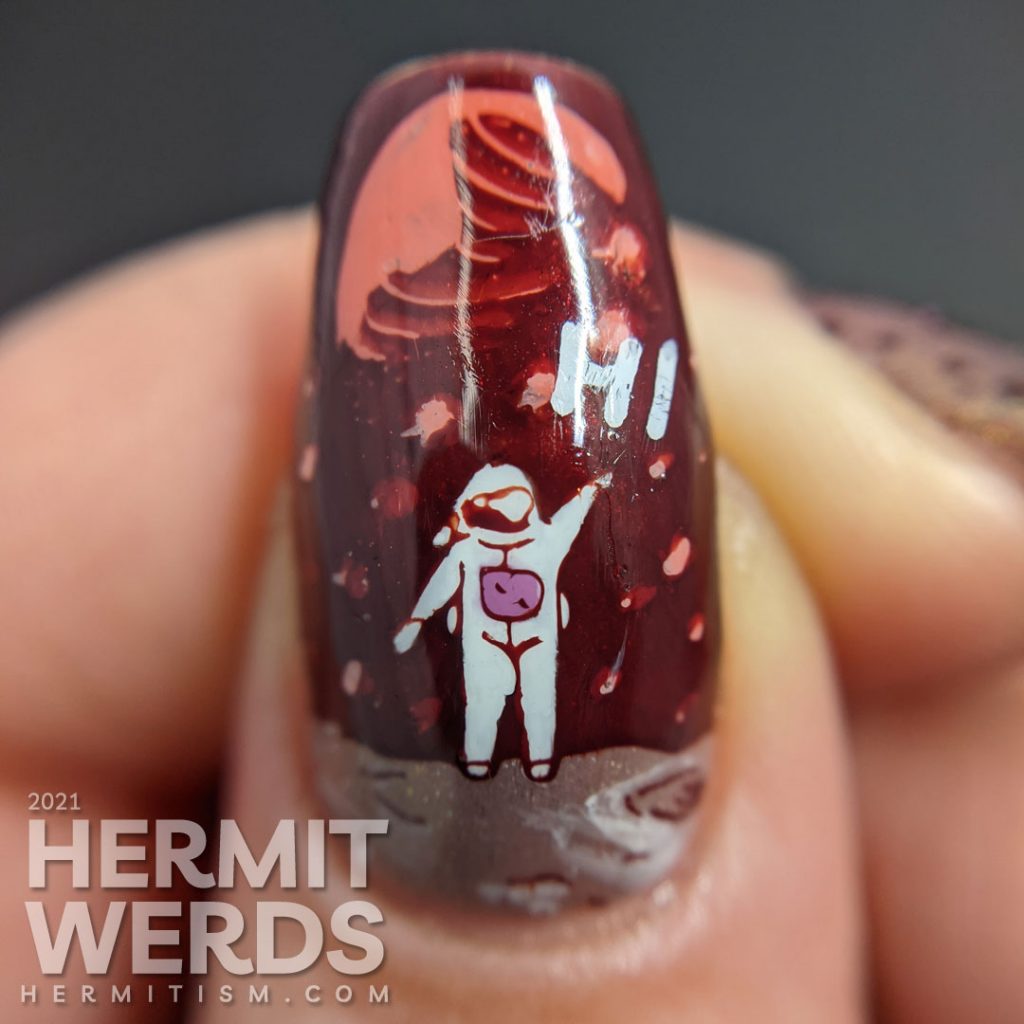 A muted red nail art with a science fiction theme. Cute aliens stamping decals scream at each other while an astronaut says "hi".