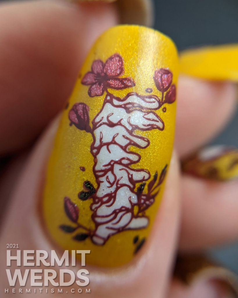 A scattered holographic mustard nail art design with tattoo-like skulls, florals, and rib cage stamping decals.