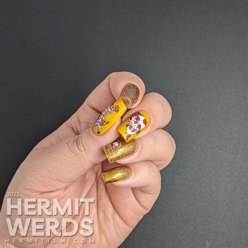 A scattered holographic mustard nail art design with tattoo-like skulls, florals, and rib cage stamping decals.