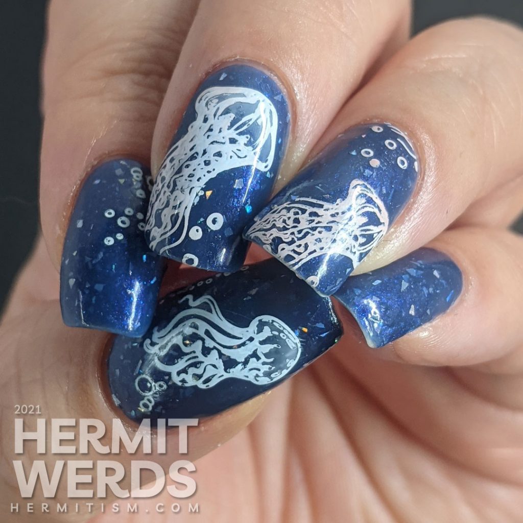 A dusty dark blue mani with transparent jellyfish stamping decals swimming in the sea with little bubbles and flashes of holo.