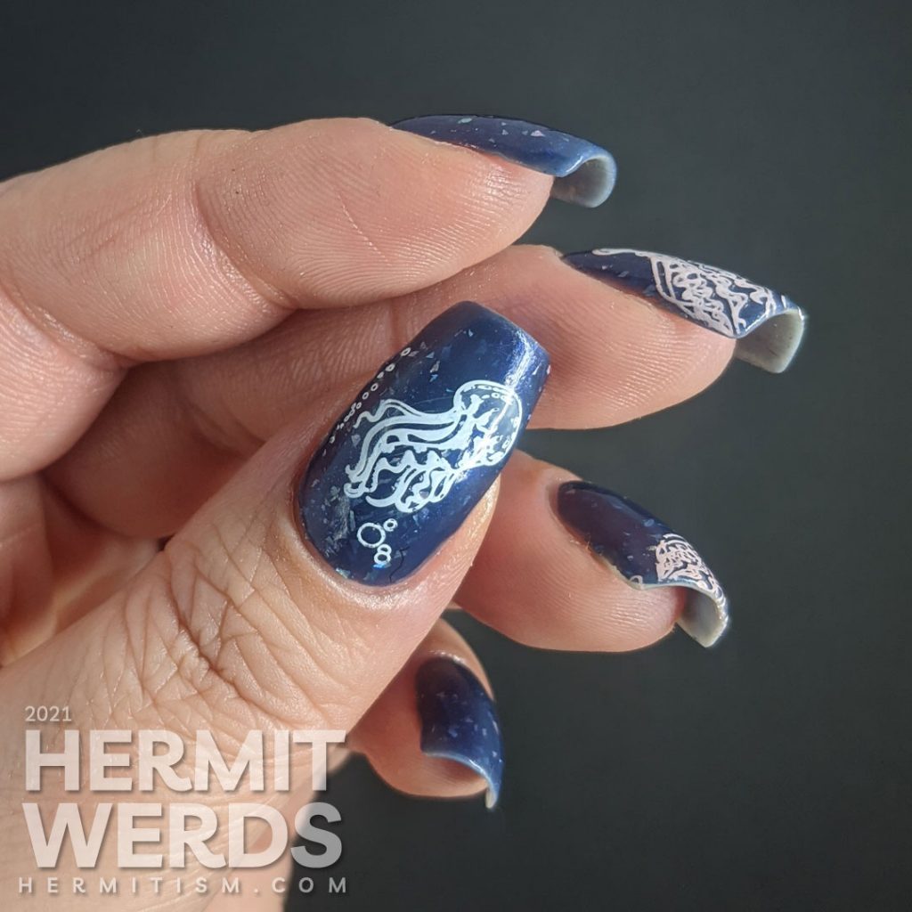 A dusty dark blue mani with transparent jellyfish stamping decals swimming in the sea with little bubbles and flashes of holo.