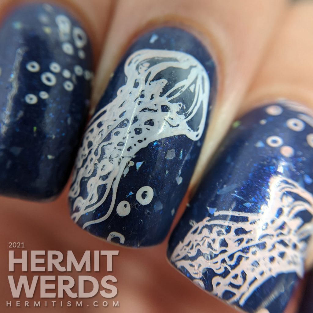 A dusty dark blue mani with transparent jellyfish stamping decals swimming in the sea with little bubbles and flashes of holo.