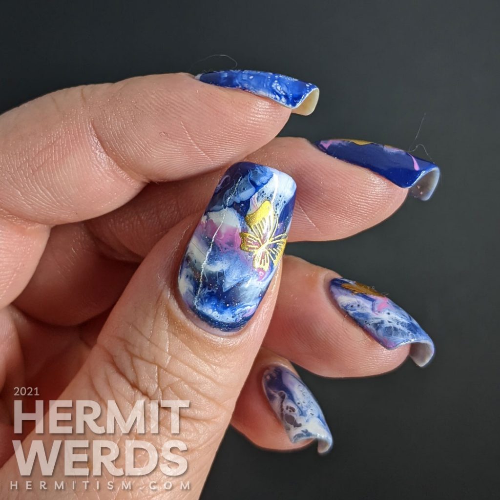 A fluid art nail art decorated with holographic gold butterfly stickers in blue, blurple, lavender, and white.