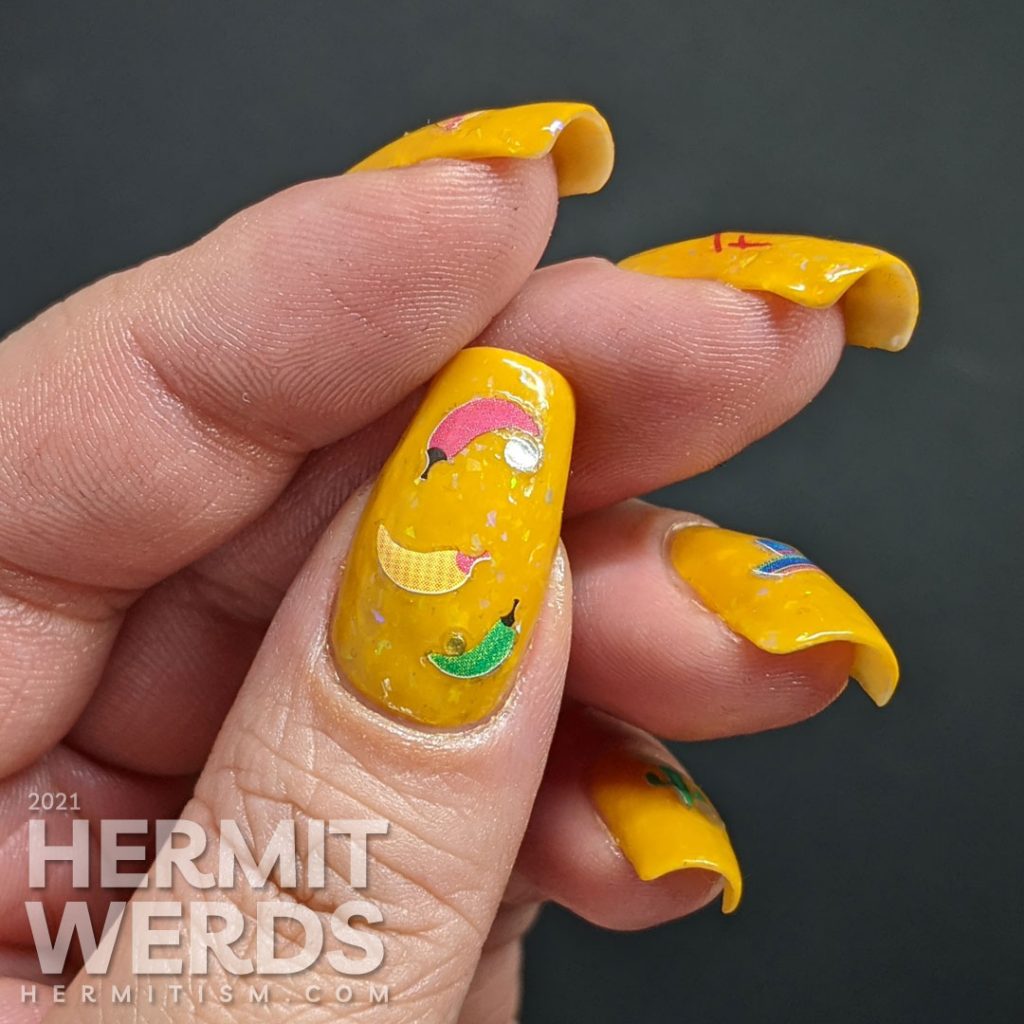 Mexico-related nail stickers (hot peppers, sombreros, maracas, and a cactus) with the word "fiesta" decorating a bright mustard crelly with iridescent flakes to celebrate Cinco de Mayo.
