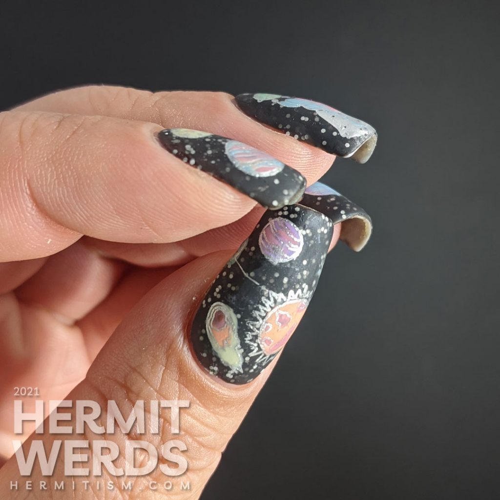 An outer space mani, with a field of stars created by white glitter in a black jelly and colorful stamping images of planets and a planet-headed lady.