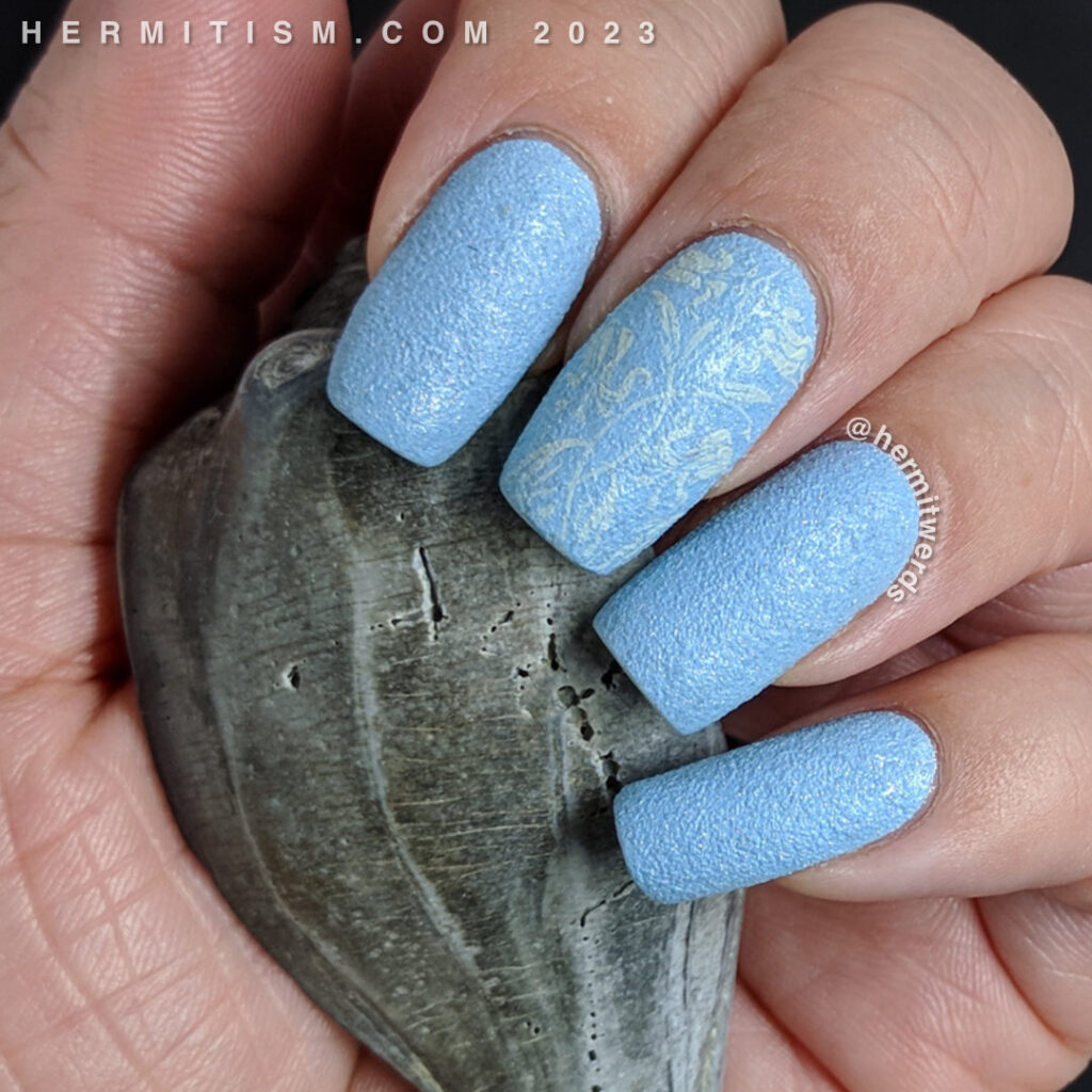 A simple light blue texture polish nail swatch with an accent nail stamped with a floral design on the texture polish.