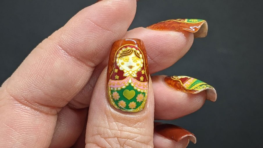 Nail art with stamping decals of beautiful Russian nesting dolls or matryoshka dolls in pinks, reds, and greens on a rusty red background with subtle jelly sandwich.