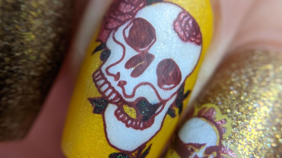 A scattered holographic mustard nail art design with tattoo-like skulls, florals, and rib cage stamping decals.