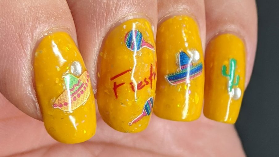 Mexico-related nail stickers (hot peppers, sombreros, maracas, and a cactus) with the word "fiesta" decorating a bright mustard crelly with iridescent flakes to celebrate Cinco de Mayo.